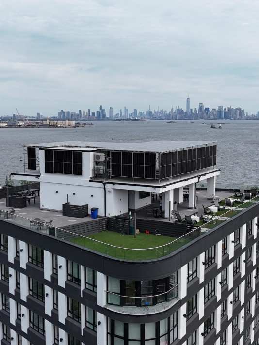 Modern building with a rooftop terrace and solar panels overlooking the Manhattan skyline across the Hudson River.