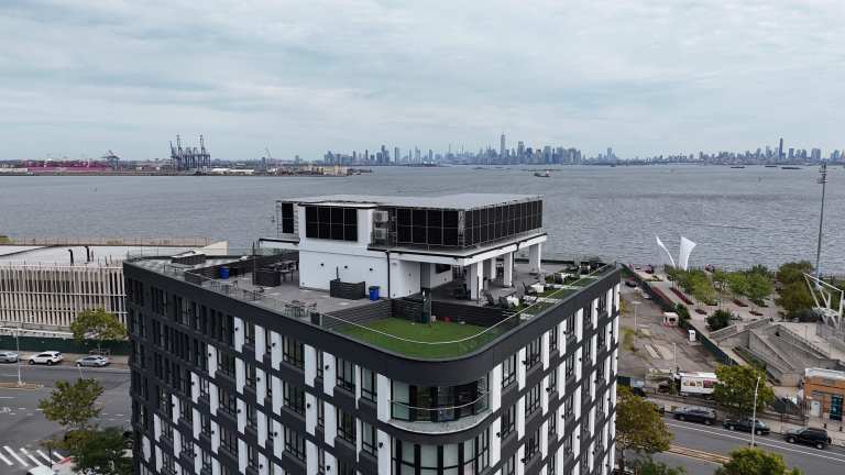 Modern building with a rooftop terrace and solar panels overlooking the Manhattan skyline across the Hudson River.