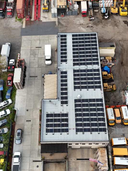 Aerial view of a commercial industrial building with rooftop solar panels surrounded by parked vehicles, equipment, and fleet buses.