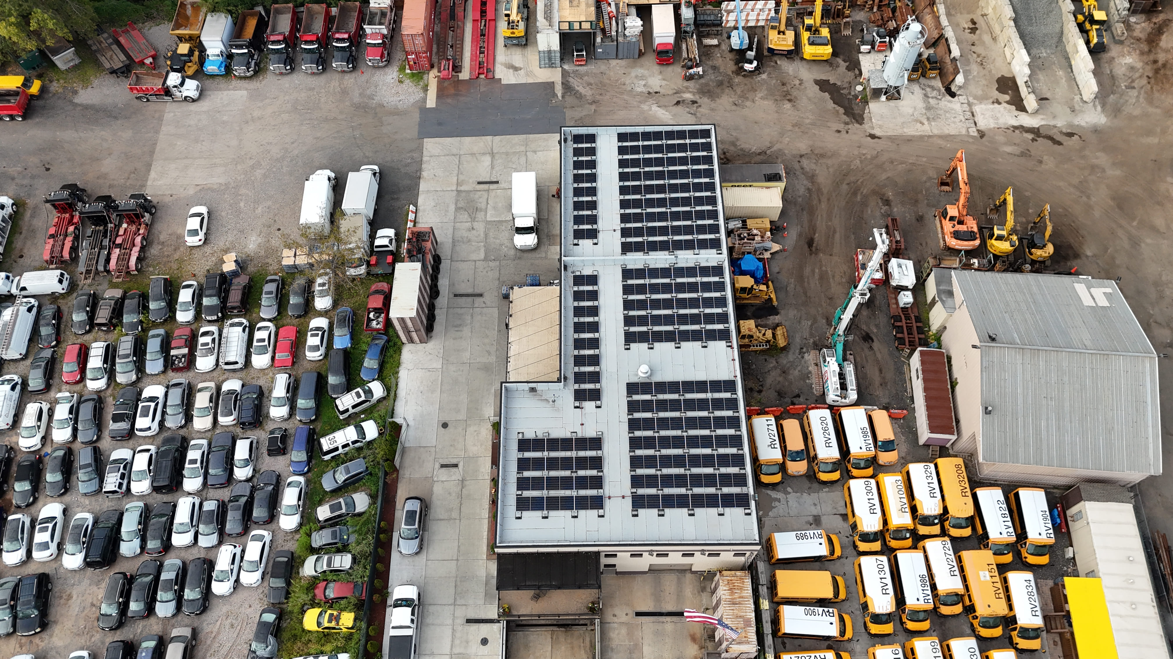 Aerial view of a commercial industrial building with rooftop solar panels surrounded by parked vehicles, equipment, and fleet buses.