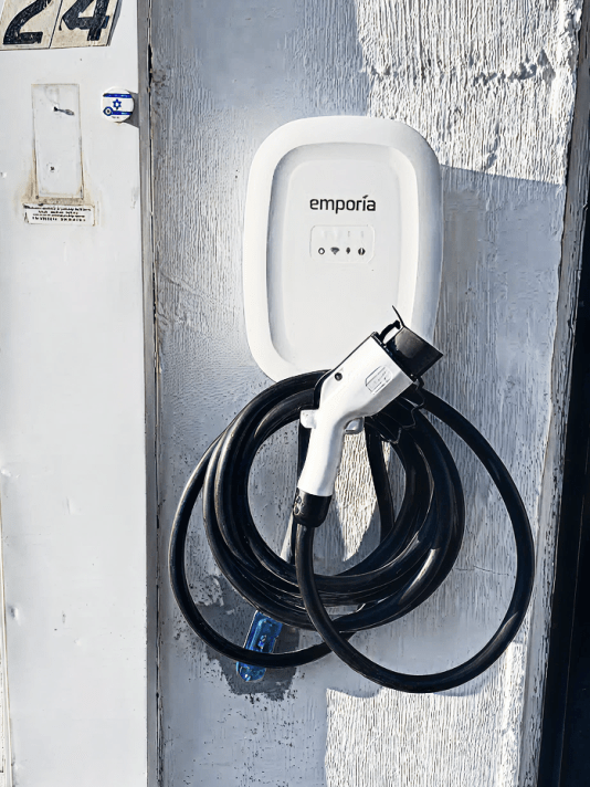 Emporia electric vehicle charger mounted on an exterior wall with a coiled charging cable and plug beside a garage door.