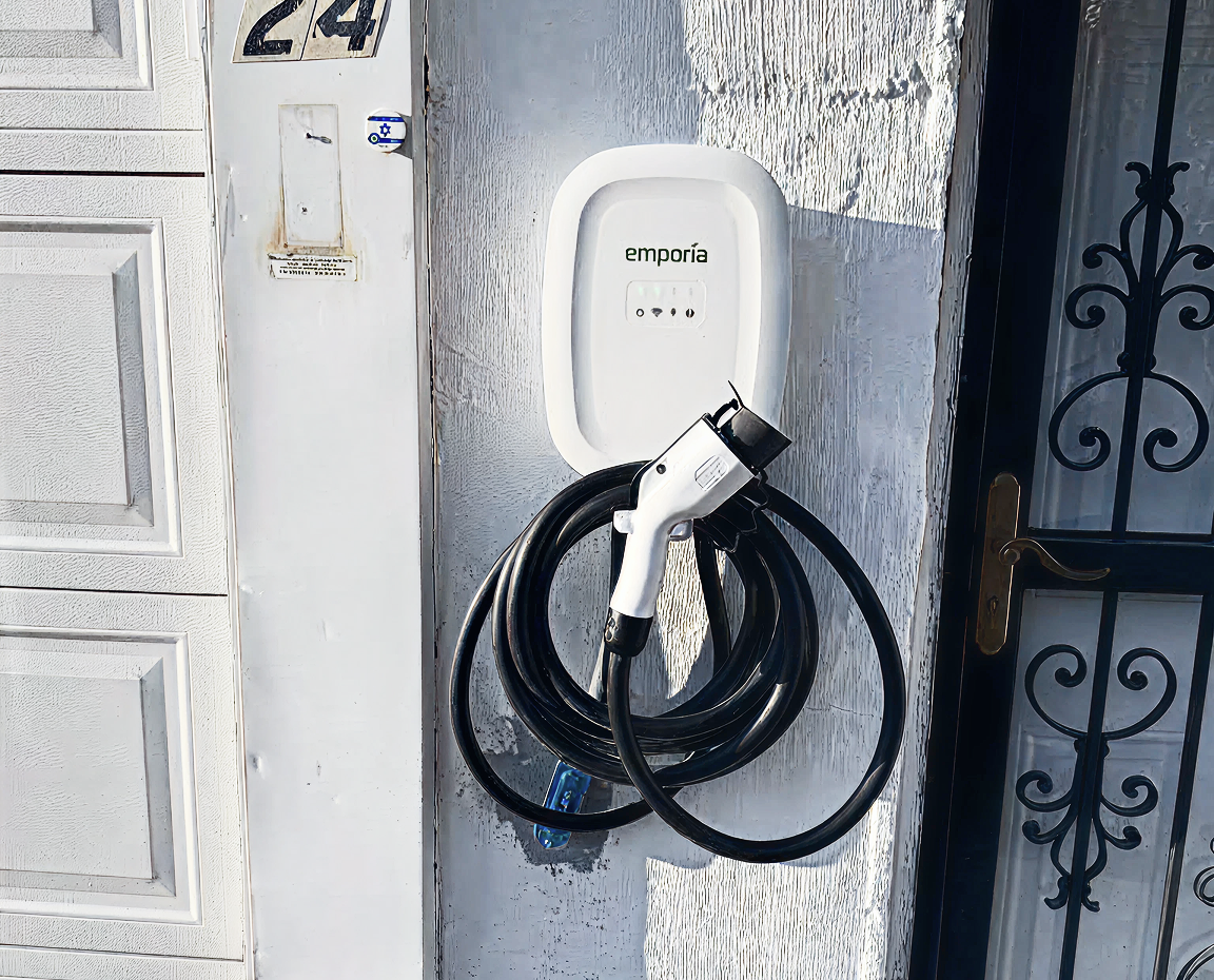 Emporia electric vehicle charger mounted on an exterior wall with a coiled charging cable and plug beside a garage door.